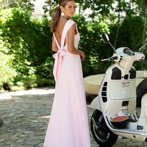 Puede incluir: Un vestido rosa p&aacute;lido sin mangas con espalda baja y lazo. Un scooter blanco est&aacute; estacionado cerca. El fondo presenta follaje verde y un camino de piedra.