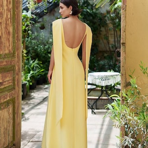 Puede incluir: Un vestido largo y fluido de color amarillo con espalda descubierta y mangas tipo capa. El vestido se lleva con tacones blancos. La imagen est&aacute; tomada al aire libre, con una puerta de madera y vegetaci&oacute;n en el fondo.
