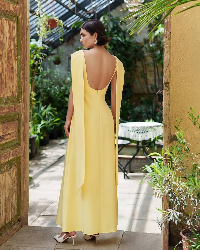 Puede incluir: Un vestido largo y fluido de color amarillo con espalda descubierta y mangas tipo capa. El vestido se lleva con tacones blancos. La imagen est&aacute; tomada al aire libre, con una puerta de madera y vegetaci&oacute;n en el fondo.