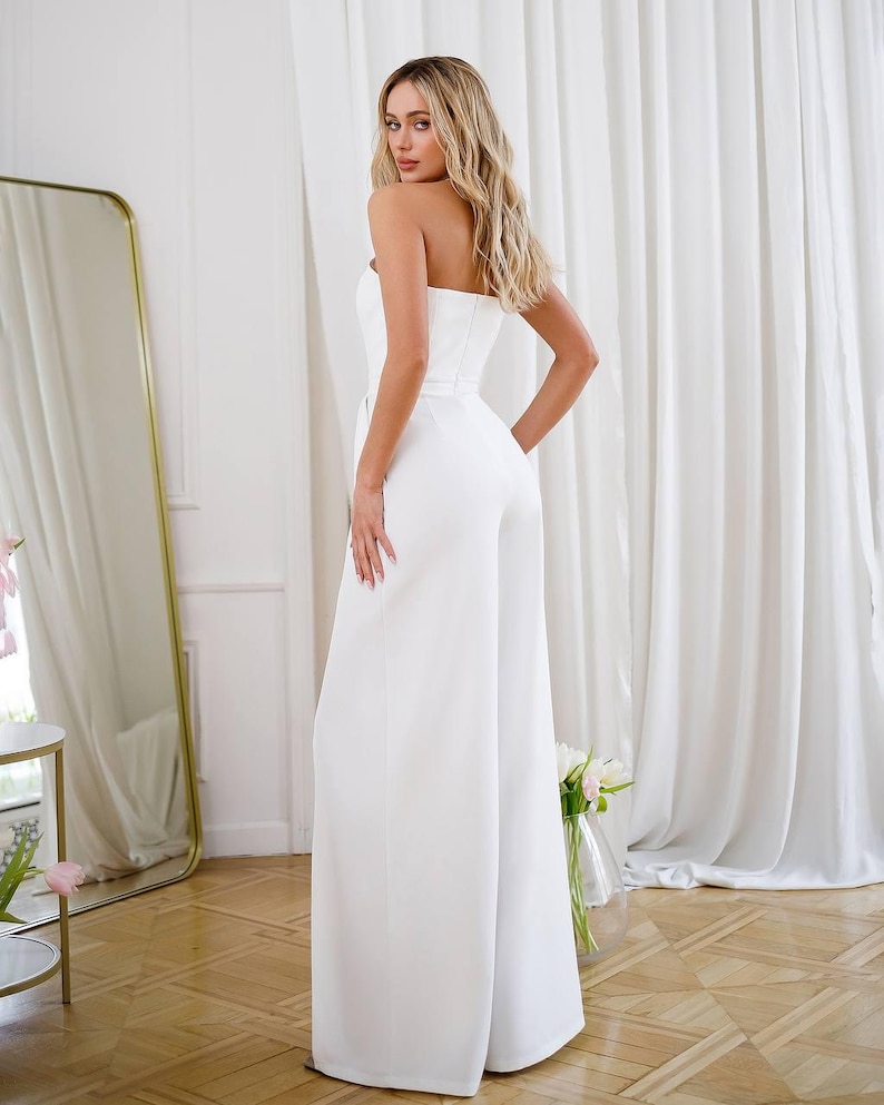 May include: A white strapless jumpsuit with a wide leg silhouette and a fitted bodice. The jumpsuit has a belt at the waist.