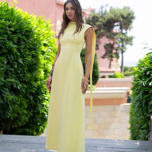 Puede incluir: Un vestido largo amarillo p&aacute;lido sin mangas, con cuello alto y falda fluida. El vestido presenta largas extensiones de tela que fluyen desde los hombros. La persona lleva pulseras doradas y tacones negros. El fondo incluye arbustos verdes y un edificio rosa.