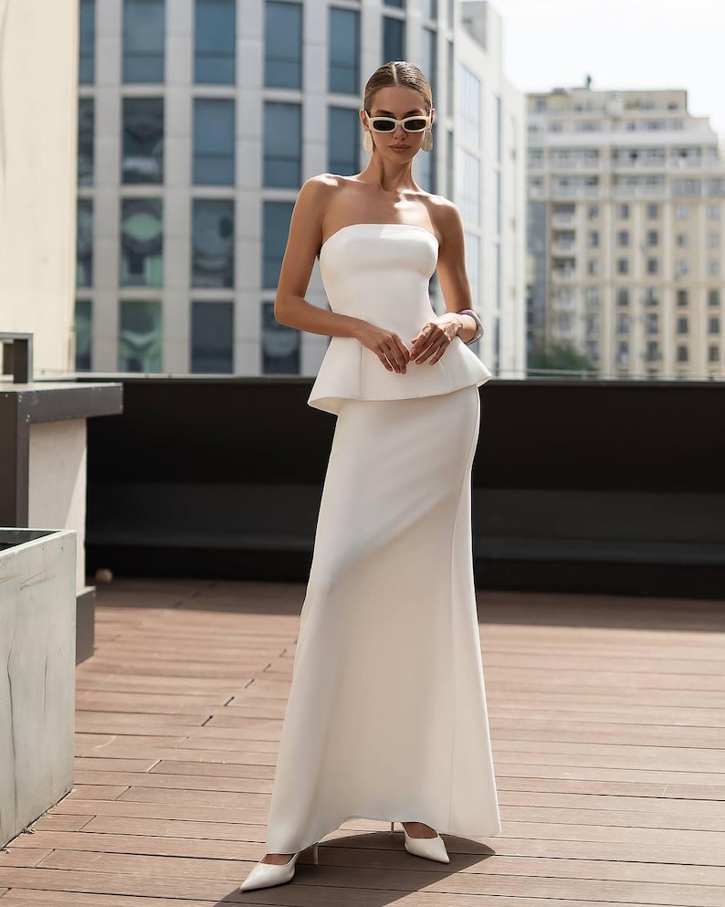 May include: A floor-length white strapless gown featuring a structured peplum waist and a fitted bodice. The dress has a smooth, solid finish and a long, flowing skirt that reaches the floor, paired with white pointed-toe heels and oval sunglasses.