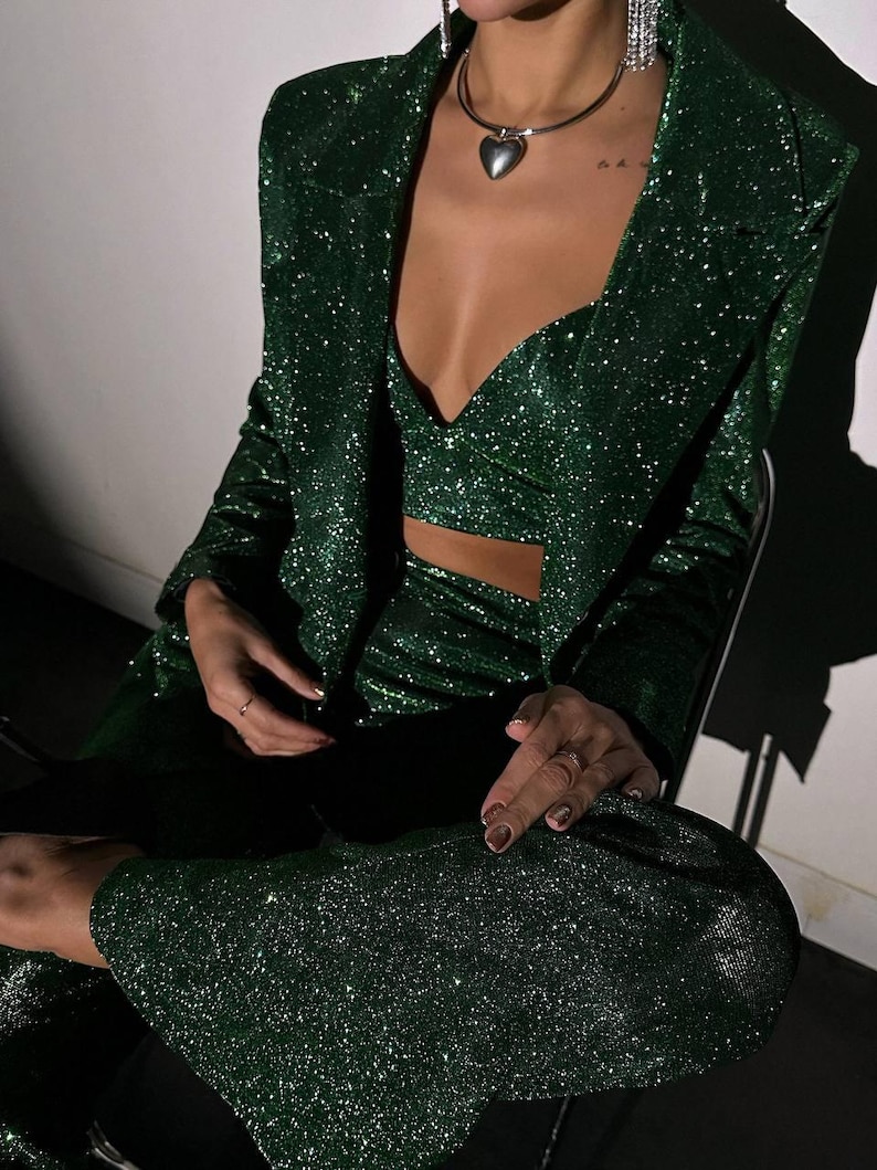 May include: A green sparkly blazer, top, and pants set. The outfit is made of a shiny fabric with a subtle glitter finish. The blazer has a classic cut with a notched lapel and long sleeves. The top is a cropped style with a sweetheart neckline. The pants are high-waisted and have a straight leg.