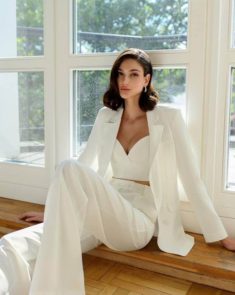 May include: A white pantsuit consisting of a blazer, crop top, and wide-leg trousers. The blazer has a two-button closure and notched lapels. The outfit is made of a smooth, possibly linen-like fabric. The model is sitting on a wooden surface.