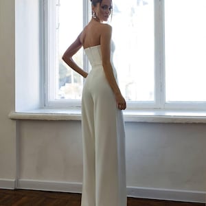 May include: A white strapless jumpsuit with a fitted bodice and wide-leg pants. The model is wearing the jumpsuit and standing in front of a window.