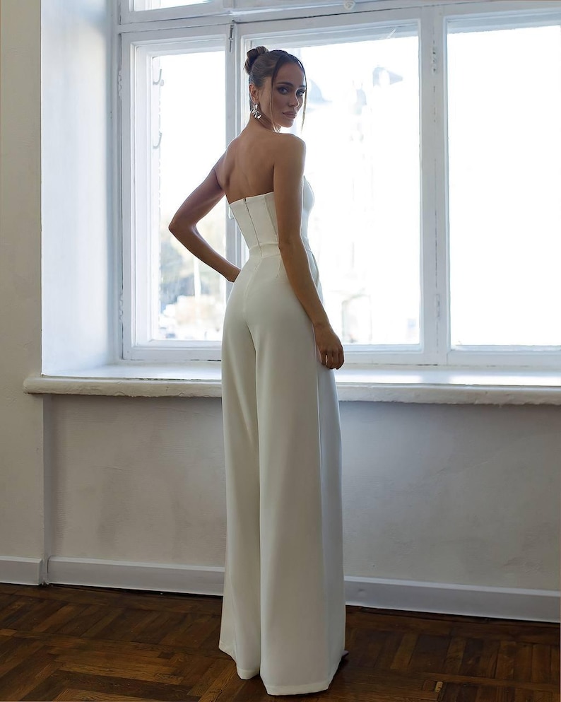 May include: A white strapless jumpsuit with a fitted bodice and wide-leg pants. The model is wearing the jumpsuit and standing in front of a window.