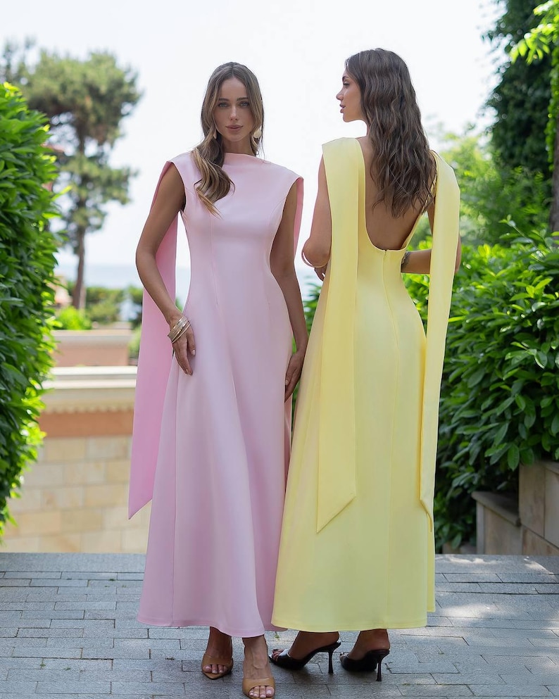 Puede incluir: Dos elegantes vestidos largos. Uno es rosa p&aacute;lido con cuello alto y mangas tipo capa. El otro es amarillo p&aacute;lido con espalda baja y mangas capa fluidas. Ambos vestidos son usados por personas de pie en escalones de piedra al aire libre.