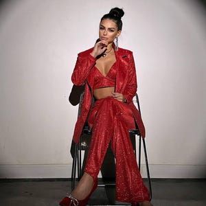 May include: A woman wearing a red sparkly blazer and pants set with a matching red top. She is sitting on a chair with her legs crossed and wearing red heels with sparkly embellishments.