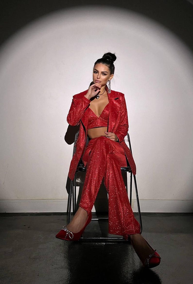 May include: A woman wearing a red sparkly blazer and pants set with a matching red top. She is sitting on a chair with her legs crossed and wearing red heels with sparkly embellishments.