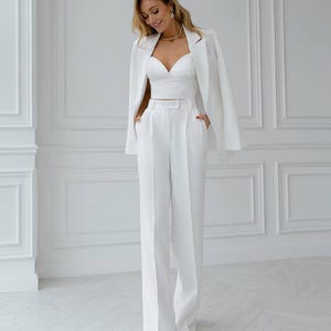 May include: A white three-piece suit consisting of a blazer, a bustier top, and wide-leg trousers. The blazer is draped over the shoulders. The outfit is accessorized with a gold necklace and earrings. The suit is made of a smooth, wrinkle-resistant fabric.
