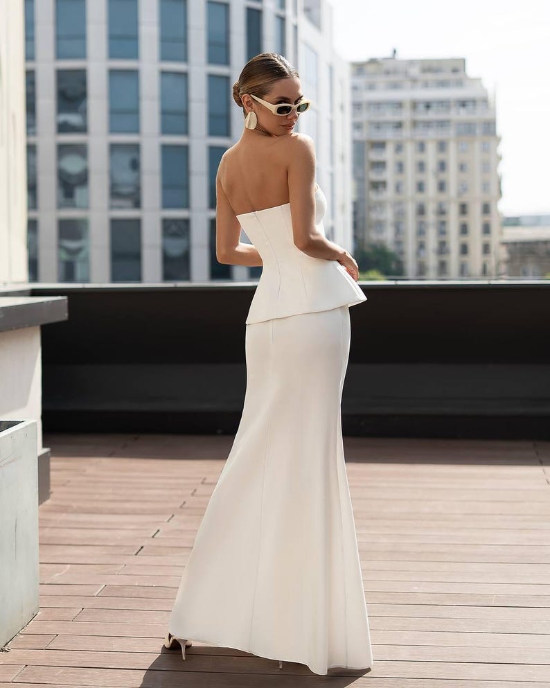 May include: A floor-length white strapless gown featuring a structured peplum waist and a fitted bodice. The dress has a clean, solid white finish and a long, flowing skirt that extends to the floor, creating a sleek and continuous silhouette.