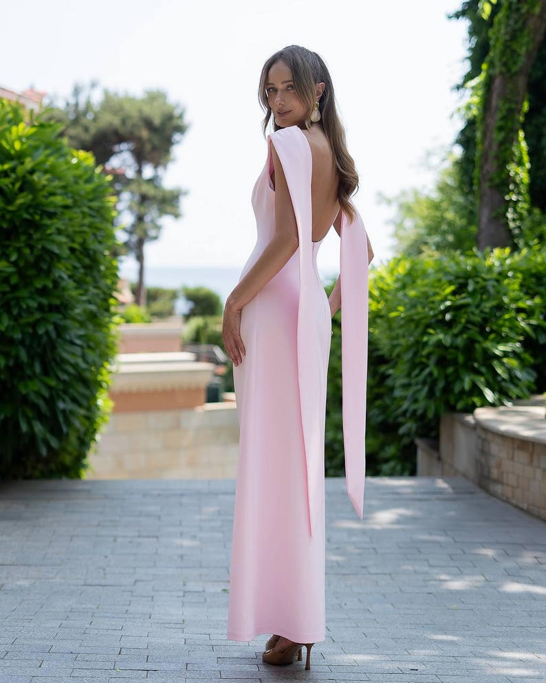 Puede incluir: Un vestido largo rosa claro, con espalda descubierta y mangas tipo capa. El vestido es entallado y est&aacute; hecho de una tela suave. La modelo lleva tacones marrones. El fondo presenta vegetaci&oacute;n y una pasarela de piedra.