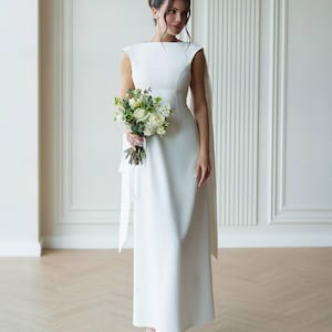 Puede incluir: Vestido de novia blanco, largo hasta el suelo, con escote barco y detalle de capa fluida. La novia sostiene un ramo de flores blancas y verdes. Lleva tacones blancos. El fondo es una pared clara con paneles verticales.