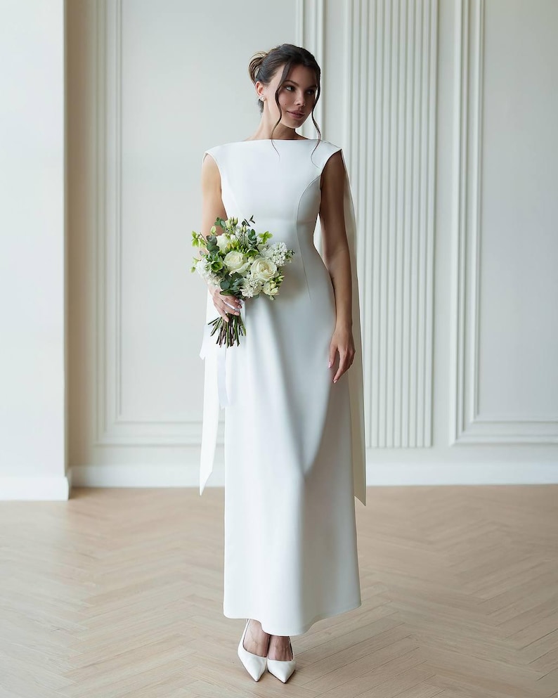 Puede incluir: Vestido de novia blanco, largo hasta el suelo, con escote barco y detalle de capa fluida. La novia sostiene un ramo de flores blancas y verdes. Lleva tacones blancos. El fondo es una pared clara con paneles verticales.