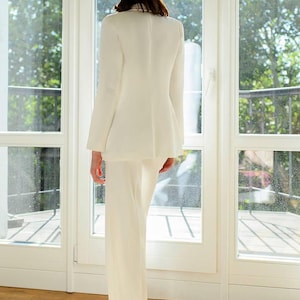 May include: A cream-colored suit, consisting of a blazer and wide-leg pants. The blazer has a tailored fit, and the pants have a high waist. The outfit is made of a smooth fabric. The person is standing in front of a large window.
