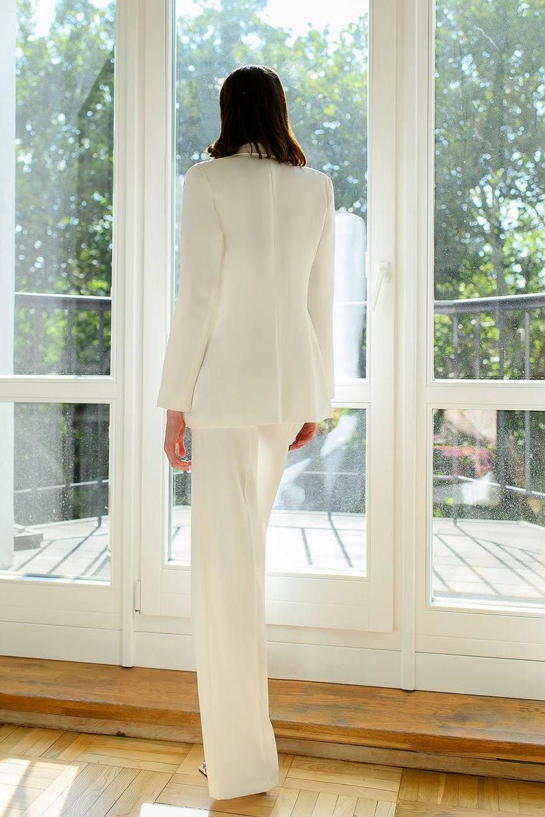 May include: A cream-colored suit, consisting of a blazer and wide-leg pants. The blazer has a tailored fit, and the pants have a high waist. The outfit is made of a smooth fabric. The person is standing in front of a large window.