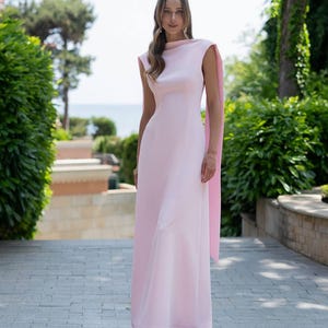 Puede incluir: Un vestido largo rosa claro con cuello alto y detalle de capa. El vestido sin mangas tiene una silueta ajustada. La modelo est&aacute; de pie en un camino de piedra, con vegetaci&oacute;n en el fondo.