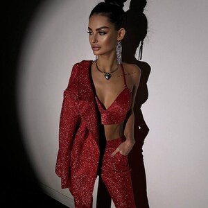 May include: A woman wearing a red sparkly pantsuit with a heart-shaped pendant necklace. The pantsuit has a blazer and flared pants.