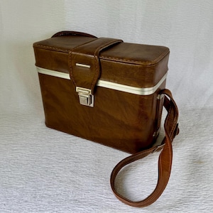 May include: A vintage brown leather camera case with a silver trim and a shoulder strap. The rectangular case has a top handle and a metal clasp closure. The leather appears worn, suggesting age and use. The case is designed to protect and carry photographic equipment.