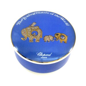 May include: A blue and gold ceramic trinket box with a design of three elephants. The box has the text "The Leading Hotels of the World" and "Chopard GENEVE" on it.