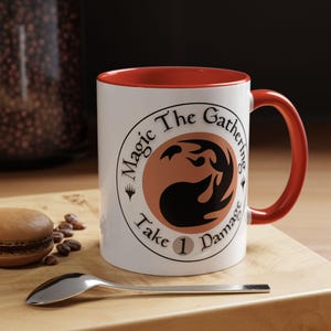 May include: White ceramic mug with a red interior and handle. The mug features the text "Magic The Gathering" in a circular design, with a stylized symbol and the words "Take 1 Damage". A spoon and a macaron are on a wooden surface.