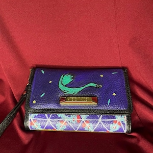 May include: A Steve Madden tri-fold wallet featuring a purple upper section with a green creature illustration and small yellow stars, and a lower section with a white geometric grid pattern over a blue and red floral print, with a black wrist strap.