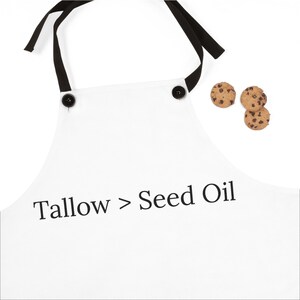 Könnte beinhalten: Weisse Schürze mit schwarzen Bindebändern und Knöpfen, mit dem Text "Tallow > Seed Oil" in Schwarz. Drei Schokoladen-Chip-Kekse befinden sich auf der rechten Seite der Schürze. Die Schürze ist für die Küche bestimmt.