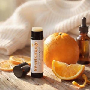 May include: A lip balm tube with the words "Sweet Orange Tallow Lip Balm" is next to an orange, orange slices, and a small brown bottle. The lip balm tube is white with black accents. The orange is whole and has a textured peel.