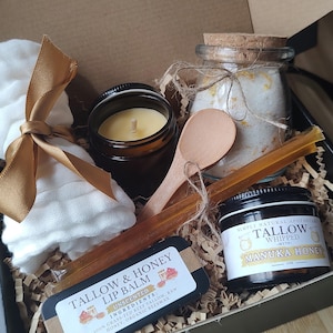 May include: A gift box filled with natural beauty products, including a jar of whipped tallow with manuka honey, a jar of bath salts, a candle, a wooden spoon, and a tin of tallow and honey lip balm. The box is lined with shredded paper and includes a white cloth.
