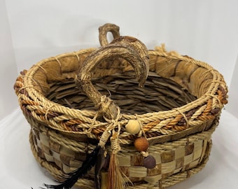 Puede incluir: Una cesta tejida con un borde de cuerda natural y un asa de cuerno de ciervo única. La cesta está decorada con plumas, cuentas y un borla.