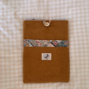 Könnte beinhalten: Eine Tablettentasche aus senfgelbem Cord mit einem floralen Stoffakzent und einem Knopfverschluss. Die Tasche hat ein kleines weißes Etikett mit dem Text "Pi & Jay" darauf.