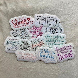 May include: A collection of glittery stickers with various inspirational quotes and phrases. The stickers feature phrases like "She is Strong," "Just Pray," and "She is Fearless." The stickers are in various colors and fonts.