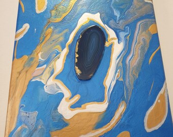 May include: Abstract canvas art with a blue, gold, and white fluid design. A dark blue stone is embedded in the center. The artwork is mounted on a white wall.