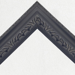 May include: A dark grey picture frame corner with decorative leaf and scroll detailing. The frame has a beveled edge and is set against a white textured background. The frame's design is symmetrical and classic.