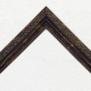 Peut inclure: Une section en forme de V d'un cadre photo. Le cadre a une finition marron foncé avec des accents dorés et un motif décoratif répétitif le long du bord intérieur. Le cadre est posé sur un fond blanc texturé.