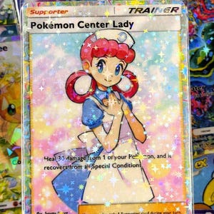 May include: A holographic Pokémon Center Lady card. The card shows a nurse with red hair and a white uniform. Text on the card includes "Pokémon Center Lady", "Trainer", and game instructions. The card is surrounded by other cards.