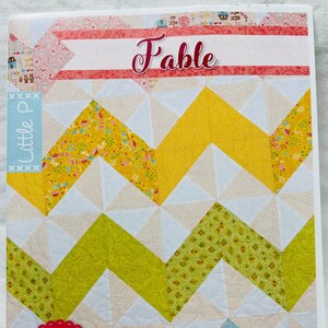 Può includere: Un modello di quilt intitolato "Fable" con un design a chevron in tessuti gialli, verdi e bianchi. Il modello è di Little P e progettato da Sarah Price. La copertina presenta un logo rosso e bianco con la scritta "it's sew emma PATTERNS".
