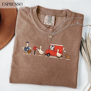 Puede incluir: Camiseta color espresso con un gráfico caprichoso de gansos repartiendo correo. El diseño incluye un ganso con uniforme postal, una furgoneta roja y un perro pequeño. Un collar plateado con un colgante está sobre la camiseta.