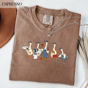 Peut inclure: T-shirt Comfort Colors couleur expresso avec un motif de cinq oies de dessin animé habillées en plombiers, chacune tenant des outils. Le t-shirt a un col rond et des manches courtes. Un collier en argent avec un pendentif est drapé sur le t-shirt.