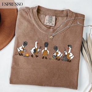 May include: An espresso-coloured t-shirt featuring a graphic of five white geese wearing brown aprons and welding helmets. The geese are depicted performing various welding tasks with tools and sparks. The shirt has a Comfort Colors brand label.