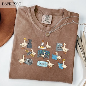 Puede incluir: Camiseta Comfort Colors color espresso con un diseño caprichoso de patos de dibujos animados interactuando con elementos temáticos espaciales como cohetes, satélites y paneles de control. La camiseta incluye el texto "Comfort Colors" y una etiqueta de talla.