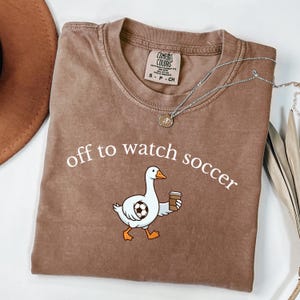 以下が含まれることがあります： 「off to watch soccer」の文字と、サッカーボールとコーヒーカップを持ったガチョウの漫画が描かれた茶色のTシャツ。クルーネックと半袖です。ペンダント付きのシルバーのネックレスがシャツの上にかけられています。