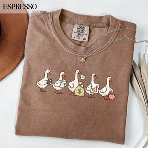 Op de afbeelding: Espresso-kleurig Comfort Colors T-shirt met een rij cartoon ganzen, elk met items zoals een rekenmachine, documenten, een geldbuidel, een grafiek en een spaarvarken. Een zilveren ketting met een hanger is over het shirt gedrapeerd.