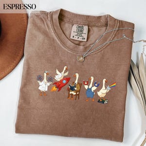 Peut inclure: T-shirt Comfort Colors couleur expresso avec un motif fantaisiste d'oies dans diverses professions, dont un scientifique, un pilote de fusée, un lecteur, une oie maniant un aimant et une oie avec un prisme arc-en-ciel. Un collier argenté est drapé sur le t-shirt.