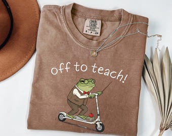 Könnte beinhalten: Ein braunes Kurzarm-T-Shirt mit einer Grafik eines Frosches, der eine Brille, eine Weste und eine rote Fliege trägt und auf einem Roller fährt. Der Text "off to teach!" ist in Weiß über dem Frosch gedruckt, der einen roten Apfel bei sich trägt.