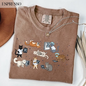 Op de afbeelding: Een bruin t-shirt met korte mouwen en een grafische print van diverse katten verkleed als medische professionals, waaronder katten met röntgenfoto's, stethoscopen en chirurgische mutsjes, verspreid tussen kleine donkerblauwe sterren.