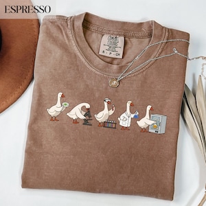 May include: Espresso-coloured t-shirt with a line of cartoon geese in lab coats and with lab equipment. The shirt has a crew neck and short sleeves. A silver necklace with a pendant is draped across the shirt.