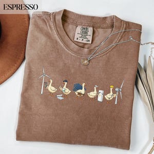 以下が含まれることがあります： エスプレッソカラーのTシャツ。風力タービンのメンテナンスやソーラーパネルの設置など、再生可能エネルギーに関連する作業を行う5羽の漫画のガチョウのグラフィックが描かれています。クルーネックで、Comfort Colorsのブランドラベルが付いています。