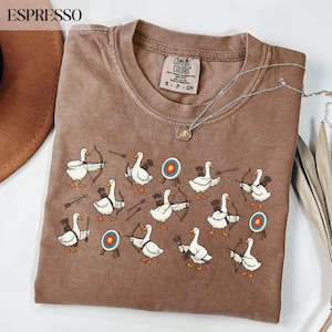 May include: Espresso-colored t-shirt with a crew neck featuring a whimsical design of white geese with bows and arrows, aiming at targets. The shirt has the text "COMFORT COLORS" on the tag. A silver necklace with a pendant is draped across the shirt.