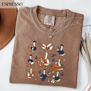 May include: Espresso-colored t-shirt with a whimsical graphic of geese in navy blue uniforms, life preservers, and a rescue boat. The shirt has a crew neck and short sleeves. A silver necklace with a pendant is draped across the shirt.